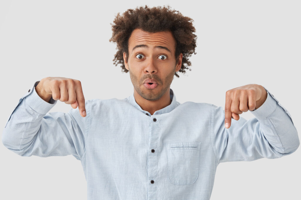 Stunned emotive young curly African American guy indicates down with both fore fingers, advertises something amazing, has surprised expression, isolated over white background. Look at floor!